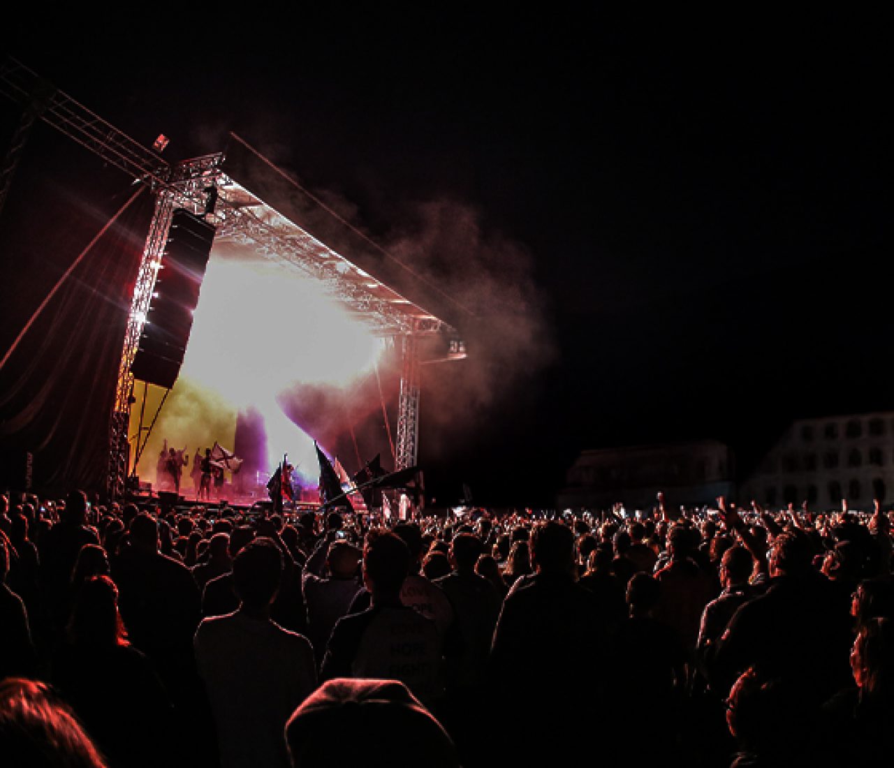 allgaeu-concerts_mood_left@2x Ein Konzert bei Nacht mit einer großen Menschenmenge vor der Bühne. Bunte Lichter und Rauch steigen von der Bühne auf, während die Menschen das Konzert genießen.