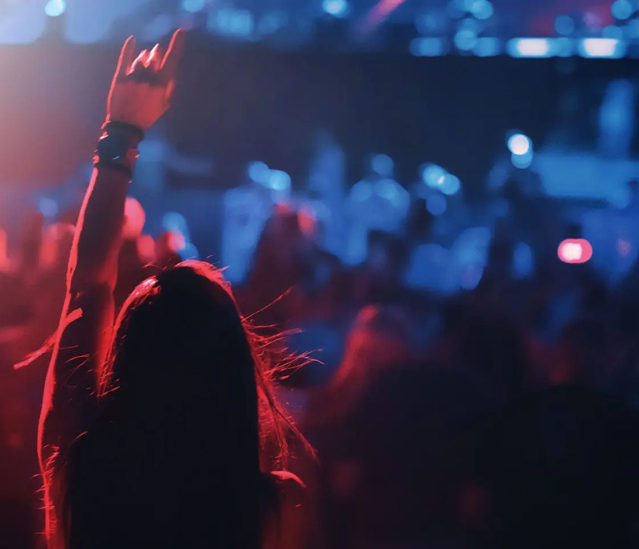 deag_mood_left@2x Eine Person steht mit erhobener Hand inmitten einer Menschenmenge auf einem Konzert oder Event. Die Szenerie ist von blauen und roten Lichtern erleuchtet, die eine energiegeladene Atmosphäre schaffen.