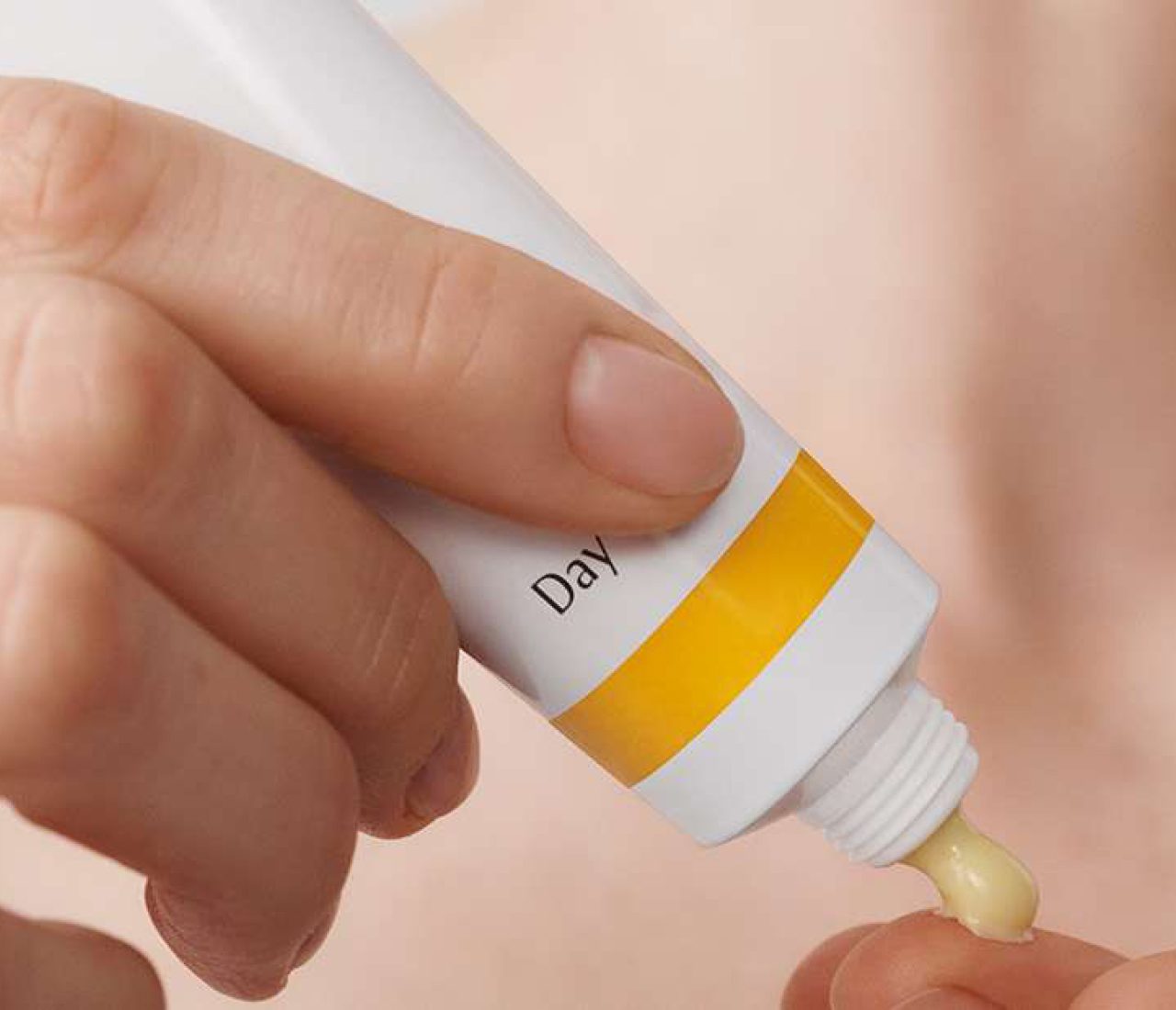 Eine Hand drückt auf eine Tube, während eine kleine Menge Creme auf die Fingerspitze herauskommt. Die Tube ist weiß mit einem gelben Streifen.
