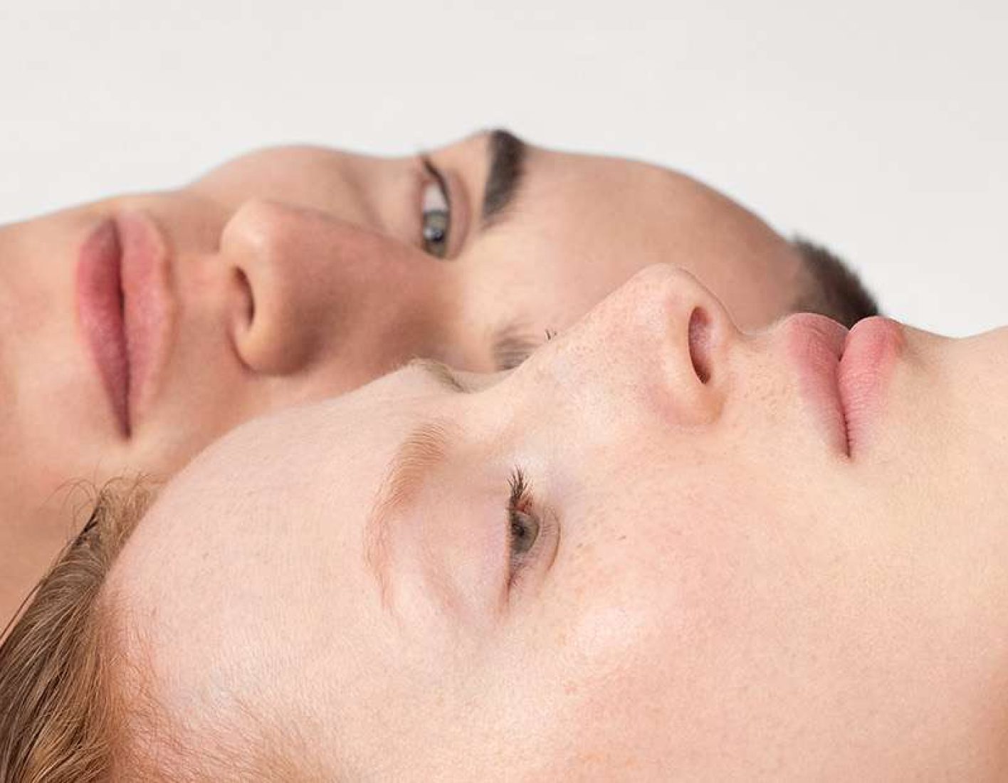 Nahaufnahme von zwei Personen, die nebeneinander auf dem Rücken liegen. Ihre Köpfe sind leicht geneigt, und ihre Augen sind geöffnet, während sie in entgegengesetzte Richtungen schauen.