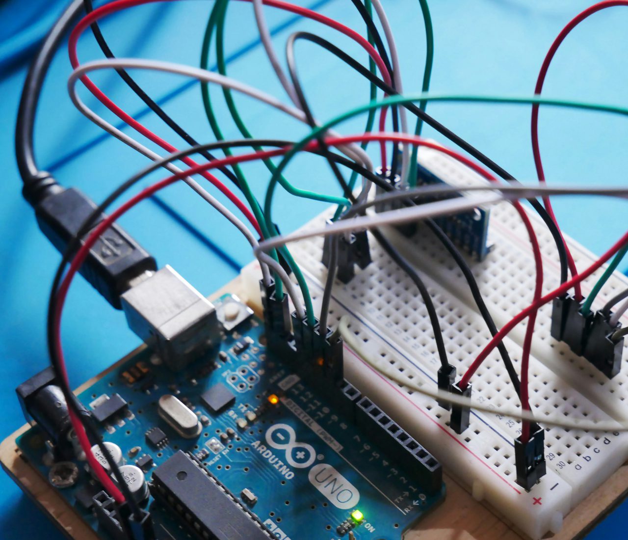 pollin_mood_left@2x Ein Arduino Uno Mikrocontroller-Board, das an ein Breadboard angeschlossen ist. Auf dem Breadboard sind mehrere Kabel verbunden, die zu verschiedenen Bauteilen führen. Das Bild zeigt eine technische Schaltung, die zur Entwicklung von Elektronikprojekten verwendet wird, wie zum Beispiel für Sensoren oder Steuerungen in DIY-Elektronik.